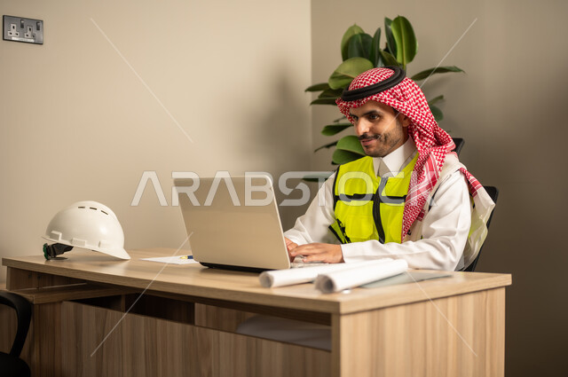 استخدام الأجهزة التقنية في المهن الهندسية، الإشراف على سير أعمال البناء، العمل في مجال الهندسة الإنشائية، متابعة تنفيذ المشاريع المعمارية، مهندس عربي خليجي سعودي يرتدي معطف الحماية يقوم بمتابعة الخطط في موقع العمل عبر جهاز اللابتوب