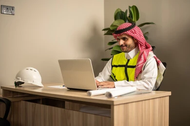 استخدام الأجهزة التقنية في المهن الهندسية، الإشراف على سير أعمال البناء، العمل في مجال الهندسة الإنشائية، متابعة تنفيذ المشاريع المعمارية، مهندس عربي خليجي سعودي يرتدي معطف الحماية يقوم بمتابعة الخطط في موقع العمل عبر جهاز اللابتوب