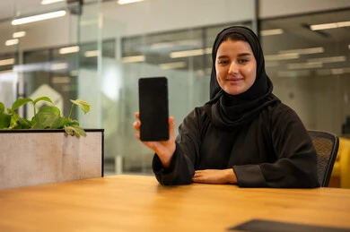 امراه عربية خليجية سعودية مبتسمة ترتدي العباءة تقوم بعرض شاشة الهاتف المحمول بإيماءات البشاشة والسرور, إنجاز المهام المكتبية في مقر الشركة، إدارة الأعمال والتواصل مع العملاء عبر الجوال,مهن ووظائف نسائية, بيئة العمل السعودية