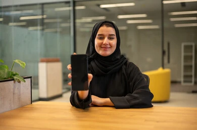 A blank black screen display, completing office tasks at the company headquarters, managing business and communicating with clients via mobile, a smiling Saudi Gulf Arab woman wearing an abaya using a mobile phone with gestures of cheerfulness and joy, women's professions and jobs, the Saudi work environment.