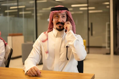 العمل في المجال المكتبي, إيماءات الجودة والرضا والقبول, رجل عربي خليجي سعودي مبتسم يرتدي الثوب التقليدي والشماغ يقوم بإجراء مكالمة هاتفية عبر الهاتف المحمول,متابعه الأعمال الادارية