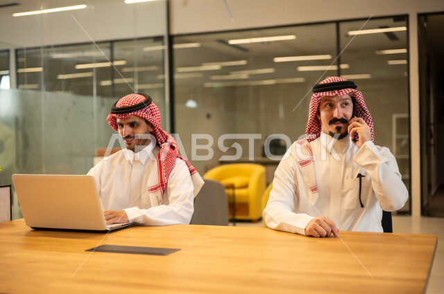 مناقشة وإنجاز مهام العمل ،دمج العمل مع التكنولوجيا المتطورة، موظفان عربيان خليجيان سعوديان يرتديان الثوب التقليد والشماغ يستخدمان الاجهزة التقنية الحديثة، مهن ووظائف سعودية، تحقيق أهداف الشركة، بيئة العمل السعودي.