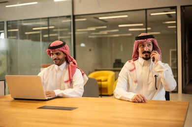 Discussion and completion of work tasks, integrating work with advanced technology, two Saudi Arabian Gulf Arab employees wearing traditional thob and ghutrah using modern technical devices, Saudi professions and jobs, achieving the company's goals, the Saudi work environment.