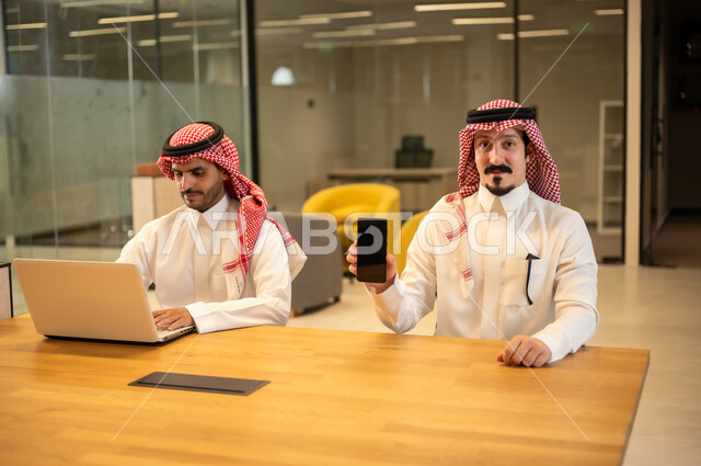 Integrating work with advanced technology, two Arab Gulf Saudi employees wearing traditional thob and ghutrah are using modern technical devices, Saudi professions and jobs, achieving the company's goals, the Saudi work environment.