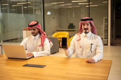 دمج العمل مع التكنولوجيا المتطورة، موظفان عربيان خليجيان سعوديان يرتديان الثوب التقليد والشماغ يستخدمان الاجهزة التقنية الحديثة، مهن ووظائف سعودية، تحقيق أهداف الشركة، بيئة العمل السعودي.