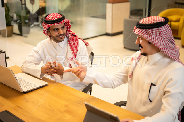 Integrating work with advanced technology, two Arab Gulf Saudi employees wearing traditional thob and ghutrah are using modern technical devices, Saudi professions and jobs, achieving the company's goals, the Saudi work environment.