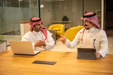 Two Saudi Arabian Gulf Arab employees wearing traditional thobes and shemaghs are using modern technological devices, discussing and completing work tasks, integrating work with advanced technology, Saudi professions and jobs, achieving company goals, and the Saudi work environment.