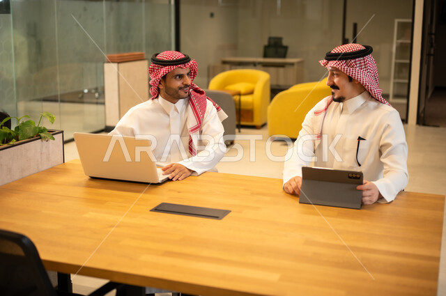 مناقشة وإنجاز مهام العمل ،دمج العمل مع التكنولوجيا المتطورة، موظفان عربيان خليجيان سعوديان يرتديان الثوب التقليد والشماغ يستخدمان الاجهزة التقنية الحديثة، مهن ووظائف سعودية، تحقيق أهداف الشركة، بيئة العمل السعودي.