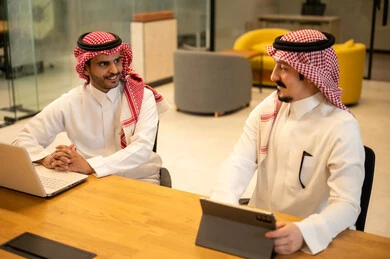 Two Saudi Arabian Gulf Arab employees wearing traditional thobes and shemaghs are using modern technological devices, discussing and completing work tasks, integrating work with advanced technology, Saudi professions and jobs, achieving company goals, and the Saudi work environment.