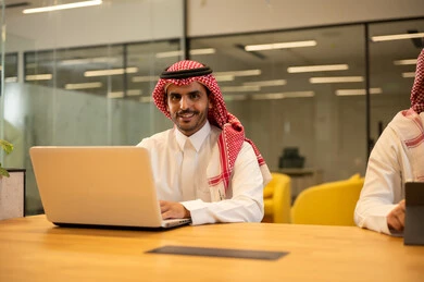 رجل عربي خليجي سعودي يرتدي الزي التقليدي والشماغ يجلس في مكتب يستخدم جهاز اللابتوب، استخدام الاجهزة التقنية الحديثة، ادارة مهام العمل اليومية، مهن ووظائف سعودية