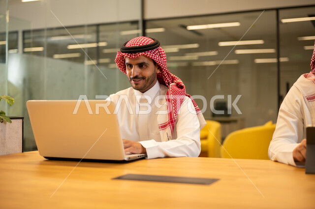 استخدام الاجهزة التقنية الحديثة، ادارة مهام العمل اليومية، رجل عربي خليجي سعودي يرتدي الزي التقليدي والشماغ يجلس في مكتب يستخدم جهاز اللابتوب، مهن ووظائف سعودية