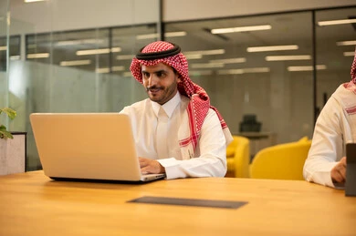 استخدام الاجهزة التقنية الحديثة، ادارة مهام العمل اليومية، رجل عربي خليجي سعودي يرتدي الزي التقليدي والشماغ يجلس في مكتب يستخدم جهاز اللابتوب، مهن ووظائف سعودية