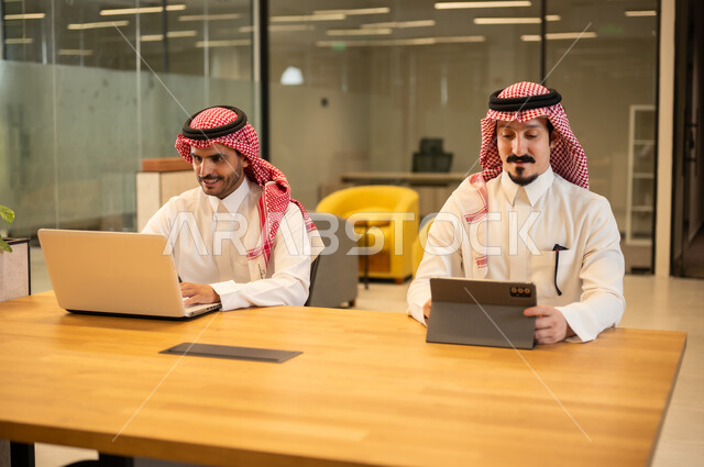 مناقشة وإنجاز مهام العمل ،دمج العمل مع التكنولوجيا المتطورة، موظفان عربيان خليجيان سعوديان يرتديان الثوب التقليد والشماغ يستخدمان الاجهزة التقنية الحديثة، مهن ووظائف سعودية، تحقيق أهداف الشركة، بيئة العمل السعودي.
