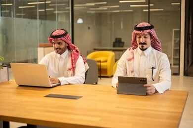 Discussion and completion of work tasks, integrating work with advanced technology, two Saudi Arabian Gulf Arab employees wearing traditional thob and ghutrah using modern technical devices, Saudi professions and jobs, achieving the company's goals, the Saudi work environment.