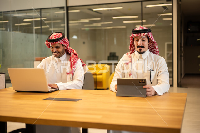 دمج العمل مع التكنولوجيا المتطورة، موظفان عربيان خليجيان سعوديان يرتديان الثوب التقليد والشماغ يستخدمان الاجهزة التقنية الحديثة، مهن ووظائف سعودية، تحقيق أهداف الشركة، بيئة العمل السعودي.