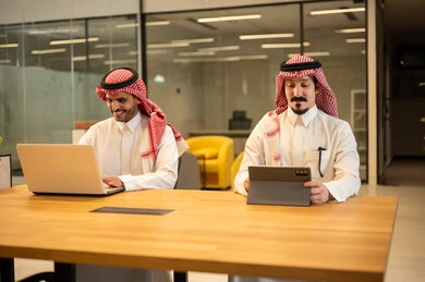 دمج العمل مع التكنولوجيا المتطورة، موظفان عربيان خليجيان سعوديان يرتديان الثوب التقليد والشماغ يستخدمان الاجهزة التقنية الحديثة، مهن ووظائف سعودية، تحقيق أهداف الشركة، بيئة العمل السعودي.