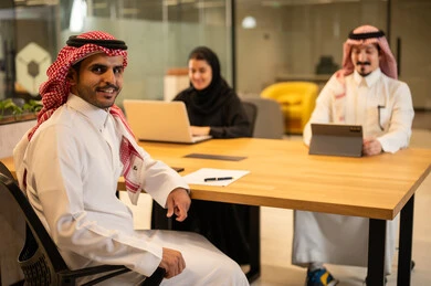 رجل عربي خليجي سعودي يرتدي الزي التقليدي والشماغ ينظر بايماءات السعادة، دمج العمل مع التكنولوجيا المتطورة، موظفين عربيين خليجيين سعوديين يستخدمون الاجهزة التقنية الحديثة، إنجاز مهام العمل ،مهن ووظائف سعودية، تحقيق أهداف الشركة، بيئة العمل السعودي.