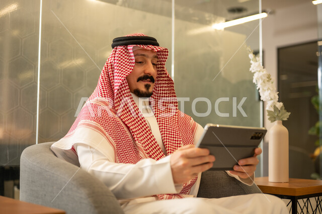 استخدام الاجهزة التقنية الحديثة، ادارة مهام العمل اليومية، رجل عربي خليجي سعودي يرتدي الزي التقليدي والشماغ يجلس في مكتب يحمل بيده جهاز التابلت، مهن ووظائف سعودية
