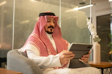 The use of modern technical devices, managing daily work tasks, an Arab Gulf Saudi man wearing traditional attire and a shemagh sitting at a desk holding a tablet, Saudi professions and jobs.