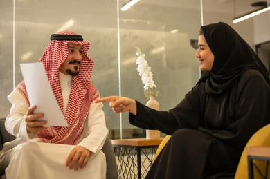 Two Saudi Arabian Gulf Arab employees are discussing work matters using papers, the concept of collaborative management to achieve the company's goals, referring to contracts and overseeing professional development, collaboration, group interaction, and problem-solving.