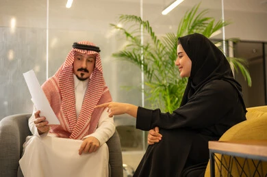 Collaboration in problem-solving, the concept of cooperative management to achieve the company's goals, two Saudi Gulf Arab employees discussing work matters using papers, referring to contracts and overseeing professional development, collaboration, collective interaction, and problem-solving.