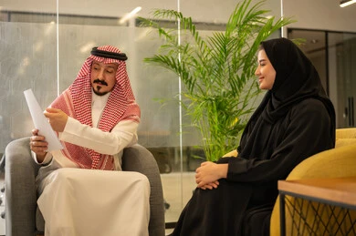 Two Saudi Arabian Gulf Arab employees are discussing work matters using papers, the concept of collaborative management to achieve the company's goals, referring to contracts and overseeing professional development, collaboration, group interaction, and problem-solving.