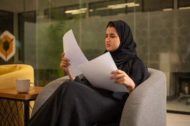مراجعة ومتابعة الخطط والمشاريع المكتبية بإيماءات التركيز، التدقيق في قراءة الملاحظات، اعداد التقارير الخاصة بالعمل، امرأة عربية سعودية خليجية محجبة ترتدي العباءة السوداء تحمل بيدها مجموعة من الاوراق البيضاء