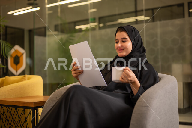 Checking notes, preparing work reports, a veiled Saudi Arabian woman wearing a black abaya holding a stack of white papers, reviewing and following up on office plans and projects with focused gestures.