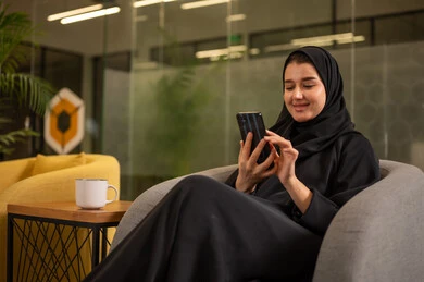 Gestures of quality, satisfaction, and acceptance, a Saudi Arabian Gulf woman wearing a black abaya is making a phone call on her mobile phone, working in the office field, following up on administrative tasks.
