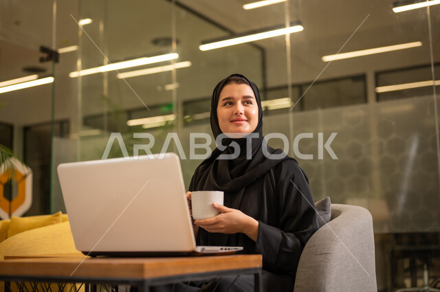Integrating work with technology and tech, providing a comfortable and suitable work environment, using a laptop to accomplish tasks professionally, an Arab Gulf Saudi woman in a hijab sitting in the office working on the laptop, employment in Saudi offices and companies.