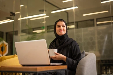 Integrating work with technology and tech, providing a comfortable and suitable work environment, using a laptop to accomplish tasks professionally, an Arab Gulf Saudi woman in a hijab sitting in the office working on the laptop, employment in Saudi offices and companies.