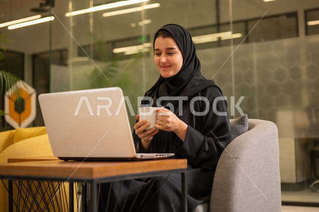Providing a comfortable and suitable work environment, integrating work with technology and tech, using a laptop to accomplish tasks professionally, an Arab Gulf Saudi woman in a hijab sitting in the office working on the laptop, employment in Saudi offices and companies.
