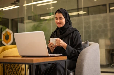 Providing a comfortable and suitable work environment, integrating work with technology and tech, using a laptop to accomplish tasks professionally, an Arab Gulf Saudi woman in a hijab sitting in the office working on the laptop, employment in Saudi offices and companies.