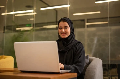 توفير بيئة عمل مريحة ومناسبة، دمج العمل مع التقنية والتكنولوجيا، استخدام جهاز الحاسوب المحمول لإنجاز المهام باحترافية، امراة عربية خليجية سعودية محجبة تجلس في المكتب تقوم بالعمل عبر اللابتوب، التوظيف في المكاتب والشركات السعودية