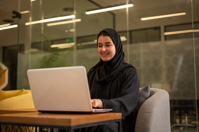 Integrating work with technology and tech, providing a comfortable and suitable work environment, using a laptop to accomplish tasks professionally, an Arab Gulf Saudi woman in a hijab sitting in the office working on the laptop, employment in Saudi offices and companies.