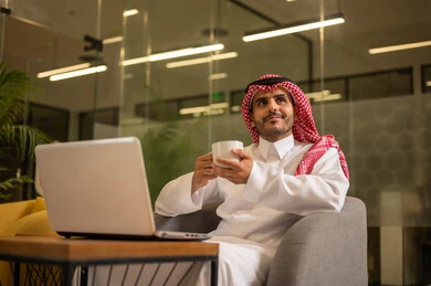 التوظيف في المكاتب والشركات السعودية، مهن وأعمال مكتبية، توفير بيئة عمل مريحة ومناسبة، استخدام جهاز الحاسوب المحمول لإنجاز المهام باحترافية، رجل عربي خليجي سعودي يرتدي الثوب والشماغ التقليدي يجلس في المكتب يقوم بالعمل عبر اللابتوب