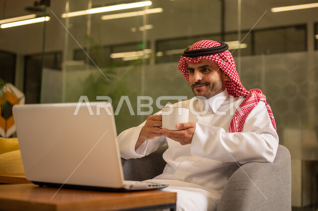 الاستمتاع بالمشروبات الساخنة، توفير بيئة عمل مريحة ومناسبة، استخدام جهاز الحاسوب المحمول لإنجاز المهام باحترافية، رجل عربي خليجي سعودي يرتدي الثوب والشماغ التقليدي يجلس في المكتب يقوم بالعمل عبر اللابتوب، التوظيف في المكاتب والشركات السعودية