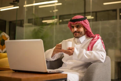 Enjoying hot beverages, providing a comfortable and suitable work environment, using a laptop to accomplish tasks professionally, an Arab Gulf Saudi man wearing traditional thobe and ghutrah sitting in the office working on the laptop, employment in Saudi offices and companies.