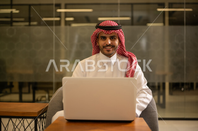 توفير بيئة عمل مريحة ومناسبة، مهن وأعمال مكتبية،استخدام جهاز الحاسوب المحمول لإنجاز المهام باحترافية، رجل عربي خليجي سعودي يرتدي الثوب والشماغ التقليدي يجلس في المكتب يقوم بالعمل عبر اللابتوب، التوظيف في المكاتب والشركات السعودية