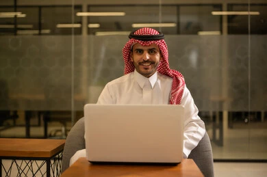 Providing a comfortable and suitable work environment, professions and office jobs, using a laptop to accomplish tasks professionally, an Arab Gulf Saudi man wearing traditional thobe and ghutrah sitting at the office working on the laptop, employment in Saudi offices and companies.