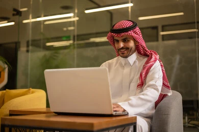 Office professions and jobs, providing a comfortable and suitable work environment, using a laptop to accomplish tasks professionally, an Arab Gulf Saudi man wearing traditional thobe and ghutrah sitting at the office working on the laptop, employment in Saudi offices and companies.