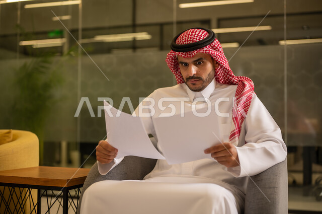 التدقيق في قراءة الملاحظات، اعداد التقارير الخاصة بالعمل،  رجل عربي خليجي سعودي يرتدي الثوب التقليدي والشماغ  يحمل بيده مجموعة من الاوراق البيضاء، مراجعة ومتابعة الخطط والمشاريع المكتبية بإيماءات التركيز