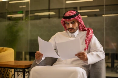التدقيق في قراءة الملاحظات، اعداد التقارير الخاصة بالعمل،  رجل عربي خليجي سعودي يرتدي الثوب التقليدي والشماغ  يحمل بيده مجموعة من الاوراق البيضاء، مراجعة ومتابعة الخطط والمشاريع المكتبية بإيماءات التركيز