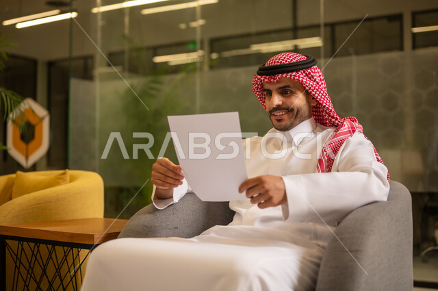اعداد التقارير الخاصة بالعمل،  التدقيق في قراءة الملاحظات، رجل عربي خليجي سعودي يرتدي الثوب التقليدي والشماغ  يحمل بيده مجموعة من الاوراق البيضاء، مراجعة ومتابعة الخطط والمشاريع المكتبية بإيماءات التركيز