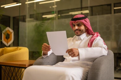 اعداد التقارير الخاصة بالعمل،  التدقيق في قراءة الملاحظات، رجل عربي خليجي سعودي يرتدي الثوب التقليدي والشماغ  يحمل بيده مجموعة من الاوراق البيضاء، مراجعة ومتابعة الخطط والمشاريع المكتبية بإيماءات التركيز