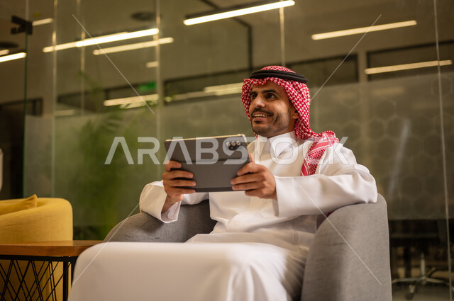 Using the tablet, managing daily work tasks, a Saudi Arabian Gulf man wearing traditional attire and a ghutrah sitting at a desk holding a tablet, Saudi professions and jobs.