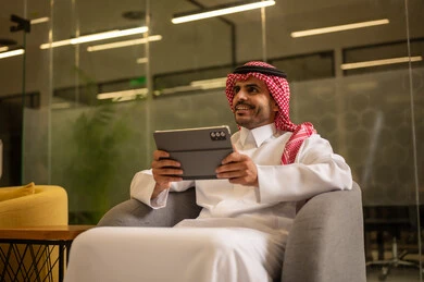 Using the tablet, managing daily work tasks, a Saudi Arabian Gulf man wearing traditional attire and a ghutrah sitting at a desk holding a tablet, Saudi professions and jobs.