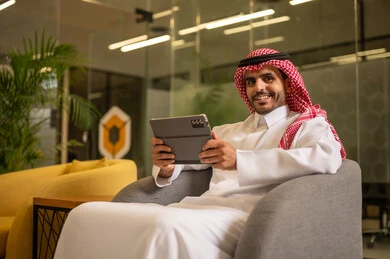مهن ووظائف سعودية، استخدام الاجهزة التقنية الحديثة، ادارة مهام العمل اليومية، رجل عربي خليجي سعودي يرتدي الزي التقليدي والشماغ يجلس في مكتب يحمل بيده جهاز التابلت