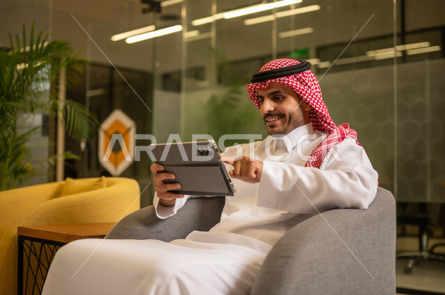 Managing daily work tasks, using modern technical devices, a Gulf Arab Saudi man wearing traditional attire and a ghutrah sitting at a desk holding a tablet, Saudi professions and jobs.