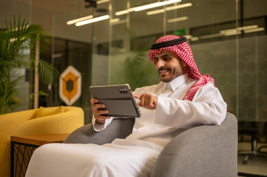 Managing daily work tasks, using modern technical devices, a Gulf Arab Saudi man wearing traditional attire and a ghutrah sitting at a desk holding a tablet, Saudi professions and jobs.